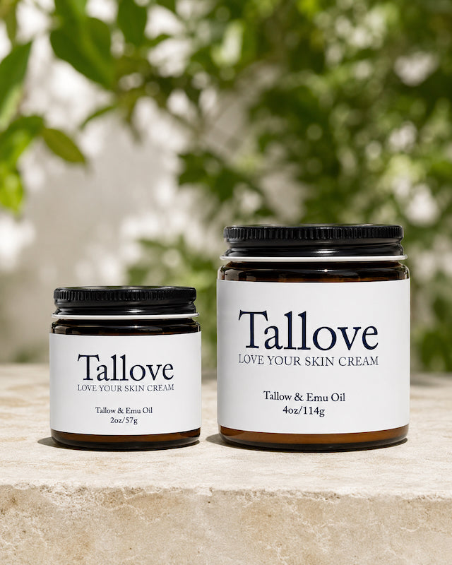 Two amber glass jars of Tallove Love Your Skin Cream displayed side by side outdoors, showing the 2 oz and 4 oz Tallow & Emu Oil sizes with clean white labels and black lids against a soft natural green background.
